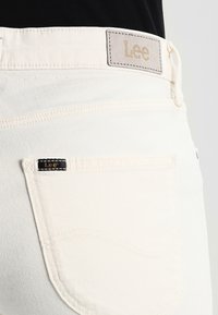 Des jeans en coton blanc présentent une étiquette en cuir avec "Lee" embossé en doré, et une petite étiquette noire à l'arrière de la poche. Texture lisse, coupe ajustée.