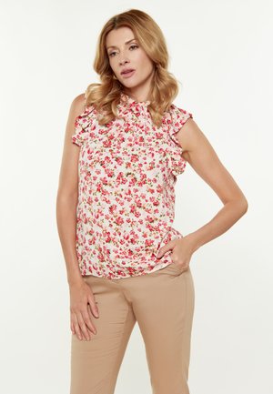Blusa floreale con maniche a volant, con uno sfondo rosa e piccoli fiori rossi e verdi. Abbinata a pantaloni beige, mani nelle tasche.