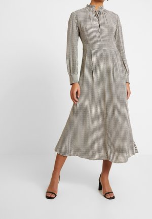 Femme portant une robe midi à manches longues, col montant, à carreaux avec plis, associée à des sandales à talons ouvertes noires sur un fond uni.