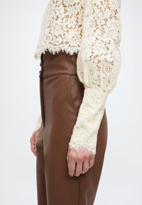 Πλαϊνή όψη ατόμου που φοράει καφέ εφαρμοστικό δερμάτινο παντελόνι και κρεμ μακρυμάνικο μπλουζάκι από δαντέλα με λουλούδια και κυματιστές άκρες.