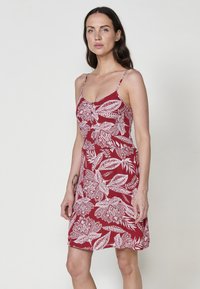 Rotes Kleid mit weißem Blumenmuster, dünnen Trägern und tailliertem Oberteil. Das Material wirkt leicht und hat eine glatte Textur.