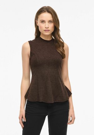 Kvinde med langt brunt hår iført ærmeløs mørkebrun peplumtop og sorte bukser, stående mod en hvid baggrund.