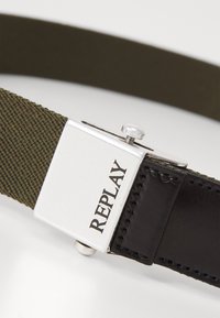 Cintura in tela verde oliva con fibbia in metallo argentato che presenta il nome del marchio "REPLAY". Accento in pelle nera alla base della fibbia.