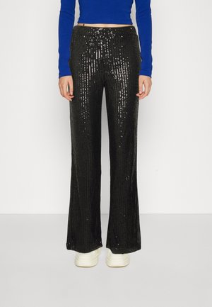 Personne portant un pantalon large à sequins noirs, un haut bleu à manches longues et court, et des baskets blanches, debout devant un fond uni.