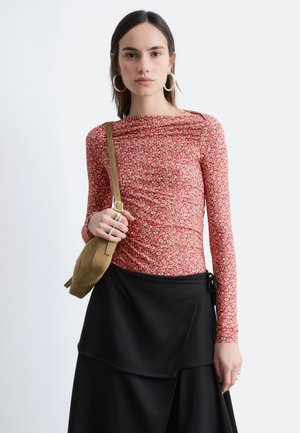 Donna con capelli lisci e scuri che indossa grandi orecchini a cerchio, top a maniche lunghe rosso con motivi floreali, gonna nera a strati e borsa a tracolla color crema, in piedi contro uno sfondo bianco.