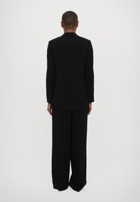 Svart skreddersydd blazer med glatt tekstur, ensidig design og matchende vide bukser. Sett fra ryggen mot en hvit bakgrunn.