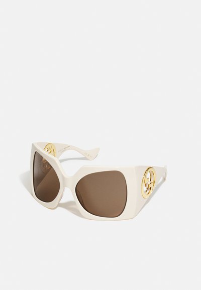 Gafas de sol blancas de gran tamaño con lentes marrones, que presentan un acento de logo dorado en la patilla y montura de plástico suave y brillante.