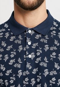 Homme portant un polo bleu marine avec un motif floral blanc et trois boutons blancs, montrant le bas de son visage et son cou avec une barbe.