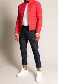 Doudoune rouge avec couture horizontale, poche zippée, chemise blanche à manches longues, pantalon sombre court, baskets blanches.