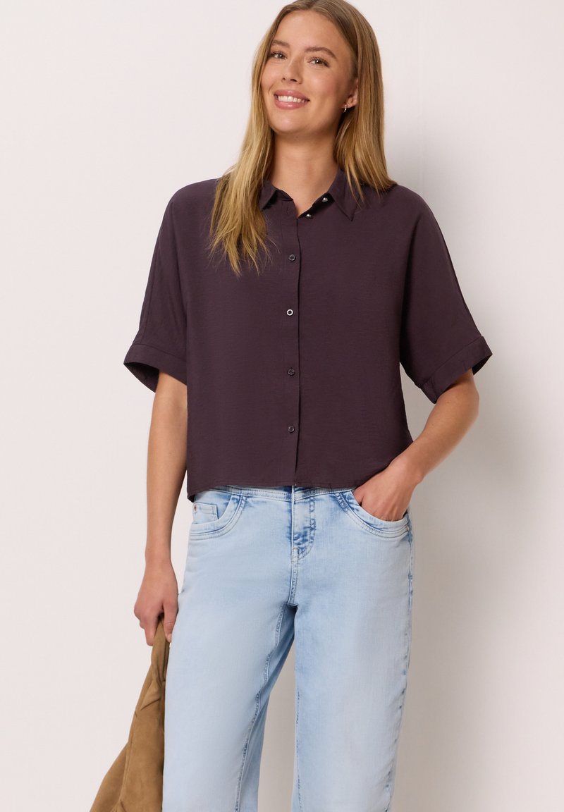 Jeune femme souriante, portant une chemise boutonnée à manches courtes violet foncé et un jean bleu clair, tenant une veste beige dans une main.