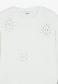 Top bianco realizzato in tessuto morbido, con intricati motivi floreali simili a pizzo nei pressi del collo e centrati sul corpo.
