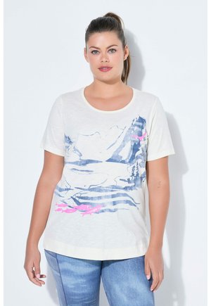 Vrouw met donker haar in paardenstaart, draagt een wit T-shirt met een blauw berg- en roze visontwerp, gecombineerd met lichtblauwe jeans, tegen een witte achtergrond.