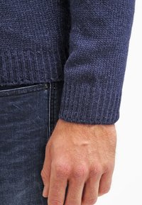 Mörk marinblå stickad tröja med ribbade muddar, i en texturerad ullblandning. Hånden visar något grov hud mot det släta tyget.