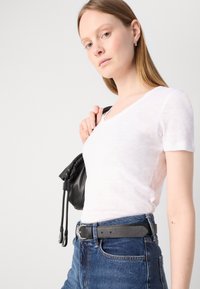 V-neck T-paita lyhyillä hihoilla valkoisena, yhdistettynä tummiin farkkuihin ja mustaan narulaukkuun. Ominaisuuksiin kuuluu kapea malli ja sileä rakenne.