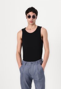 Zwart mouwloos tanktop gecombineerd met lichtgrijze plooibroek. Het model draagt ronde zonnebrillen, die een minimalistisch ontwerp tonen.