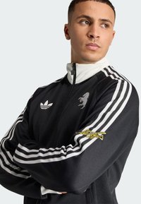 Chaqueta negra con cremallera, cuello alto, mangas a rayas blancas y negras, logo en relieve y texto bordado en amarillo en el brazo.