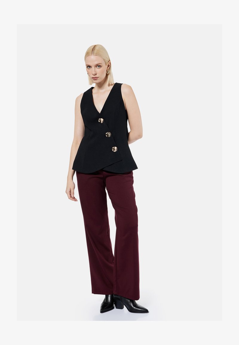 Woman wearing sleeveless black V-neck top with three large buttons and burgundy wide-leg pants and black heeled boots.
