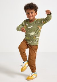 Maglietta verde a maniche lunghe con stampa di dinosauro, abbinata a pantaloni jogger marroni con dettagli testurizzati e sneakers gialle con strisce blu e bianche.