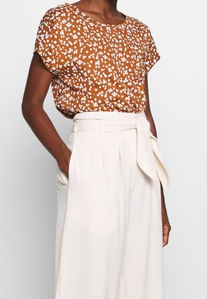 Kvinde iført en brun kortærmet top med hvidt blomsterprint, stoppet ned i højtaljede, hvide viddebukser med stofbælte.