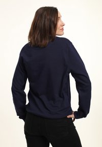 Námořnická modrá mikina s dlouhými rukávy, s límcem ve tvaru kruhu, jemná textura a boční rozparky; nošena s černými kalhotami, viděná zezadu.