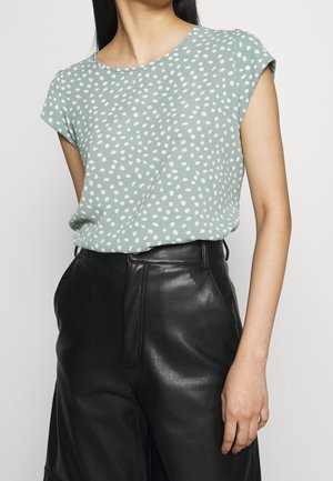 Femme portant un haut vert clair à manches courtes avec des pois blancs et un pantalon en cuir noir taille haute sur un fond uni.