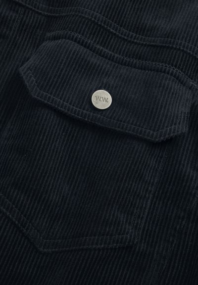 Černá kordurovná látka s vertikálními pruhy, s náprsní kapsou a kovovým logem knoflíku vyrytým "W.W." na klopě.