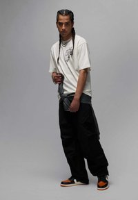 Weißes Grafik-T-Shirt, schwarze Cargo-Hose, grauer Stoff um die Taille gebunden, schwarz-orange Sneakers, mittellange geflochtene Haare, selbstbewusste Pose.