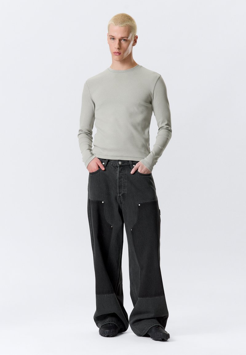 Ανοιχτό γκρι μπλουζάκι με μακριά μανίκια και υπερμεγέθη σκουρόχρωμα γκρι τζιν παντελόνια με ορατές τσέπες και ασημένιες μεταλλικές λεπτομέρειες. Το μοντέλο στέκεται με τα χέρια στις τσέπες.