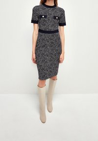 Abito tweed bianco e nero con maniche corte, caratterizzato da due tasche frontali e bottoni dorati. Abbinato a stivali beige al ginocchio.