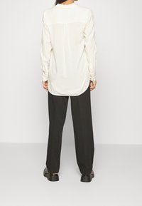 Camicia a maniche lunghe color crema con vestibilità rilassata, dettaglio della piega posteriore e polsini con bottoni, abbinata a pantaloni neri a gamba larga e scarpe nere.