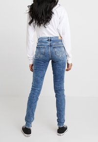 Mujer de pie de espaldas a la cámara, vistiendo jeans ajustados azul desgastado, camisa blanca de manga larga y zapatillas negras sobre fondo blanco.