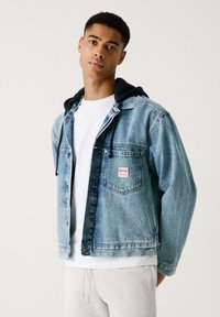 Jeansjack met een lichtblauwe wassing, voorzien van een voorkantzak, een rood label en een bevestigd marineblauwe hoodie. Gedragen over een wit t-shirt en grijze broek.