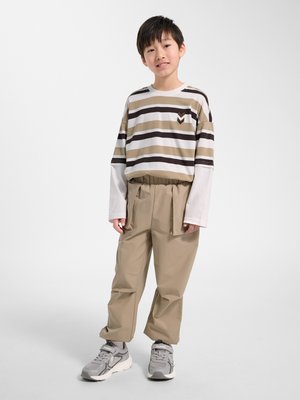 Dreng stående, iført beige og hvidstribet langærmet skjorte, beige bukser med stropper og grå sneakers på en ensfarvet hvid baggrund.