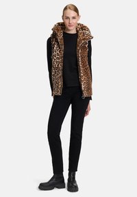 Kamizelka w panterkę z wysokim kołnierzem, miękką teksturą i zapięciem na zamek, noszona na czarnej koszuli z długim rękawem i czarnych spodniach, z masywnymi bootsami.