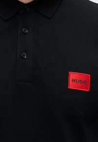 Czarna koszulka polo z trzema guzikami i czerwonym prostokątnym naszywką z napisem "HUGO" na lewej piersi.