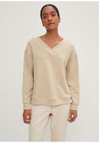 Beige sweatshirt med V-halsdesign og faldne skuldre. Fremstillet af blødt stof; har ribbede manchetter og kant. Bærer den med lyse jeans.