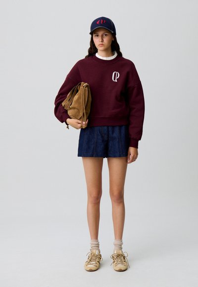 Sudadera burdeos con logotipo, pantalones cortos de mezclilla, bolso de ante marrón, gorra de béisbol oscura, zapatillas deportivas beige y calcetines claros. Texturas suaves en todo.