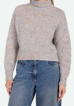Femme portant un pull à col roulé tricoté multicolore clair et un jean taille haute bleu sur un fond uni.
