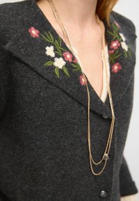 Grau gestrickte Strickjacke mit floraler Stickerei in Rosa, Weiß und Grün am Kragen. Auch eine mehrlagige goldene Kette ist sichtbar.
