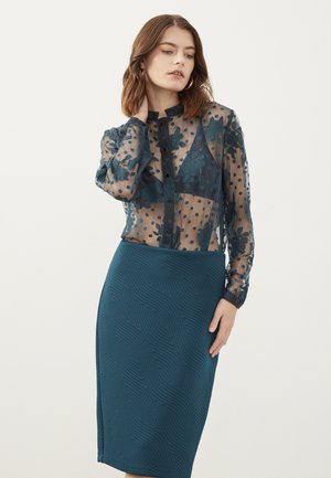 Doorzichtig, marineblauw blouse met stippen en bloemenborduursel, gecombineerd met een getextureerde teal kokerrok met een golfpatroon.