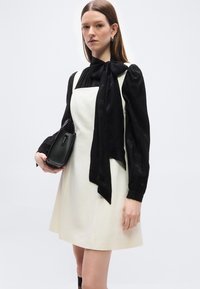 Γυναίκα που φοράει λευκό φόρεμα χωρίς μανίκια πάνω από μαύρο μπλουζάκι με μακριά μανίκια και μεγάλο φιόγκο, κρατώντας μια μαύρη τσάντα με υφή.