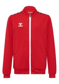 Rød træningsjakke med en tekstureret overflade, hvid lynlås, sidelommer og sort chevron mønster langs skuldrene. Hummel-logo i hvid.