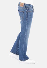 Jean de mezclilla azul con corte recto, que presenta un parche trasero de cuero marrón y un sutil detalle de costura en los bolsillos traseros.