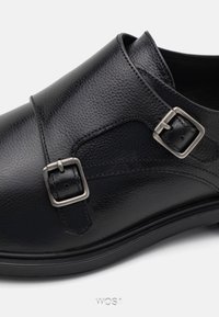 Zwarte leren schoen met een steentjesstructuur, gekruiste bandontwerp met twee metalen gespen en een lage profielzool.