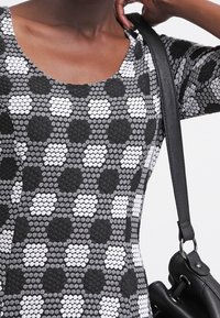 Czarna i biała sukienka wzorowana, z okrągłym dekoltem, ozdobiona heksagonalnymi kształtami i teksturowanym materiałem. Czerwona torba spoczywa na ramieniu.