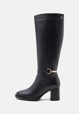 Botte noire en cuir hauteur genou avec talon bloc, sangle décorative à boucle dorée autour de la cheville, et petit logo doré près du haut.