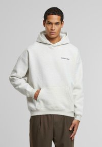 Lys grå sweatshirt med hætte, frontlomme og ribbede manchetter. Har "AnotherCotton" logo i sort på venstre brystområde.