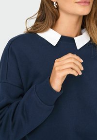 Felpa blu navy con colletto Peter Pan bianco. Presenta polsini a coste e una vestibilità ampia; texture liscia con dettagli minimi.