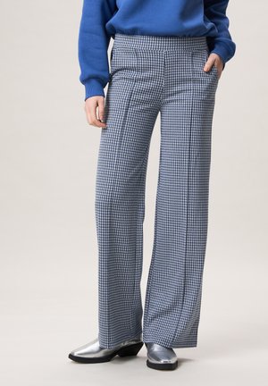 Persoon draagt blauw-witte wijde broek met hondentandpatroon, blauwe trui en zilveren laarzen met spits toelopende neus, handen in de zakken.