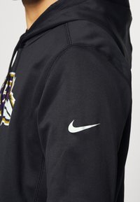 Personne portant un sweat à capuche Nike noir avec un logo swoosh blanc sur la manche et une partie d'un graphisme multicolore visible sur la poitrine.